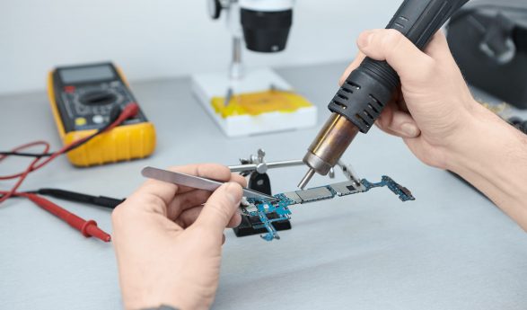 Candid shot of unrecognizable repairman soldering components in motherboard while fixing damaged smart phone, using tweezers and iron. Computer hardware, electronics, repair and upgrade concept